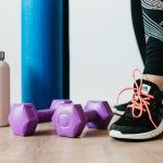 Crop sportswoman wearing sneakers and black leggings standing with sport mat on wooden floor near dumbbells and water bottle before exercising indoors