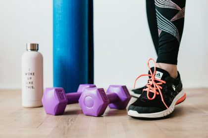 Crop sportswoman wearing sneakers and black leggings standing with sport mat on wooden floor near dumbbells and water bottle before exercising indoors